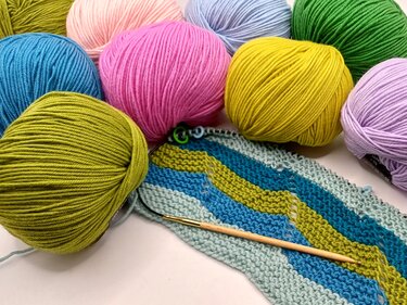 Nahaufnahme bunter Wollknäuel neben gestreiftem Strickstück und Rundstricknadel.