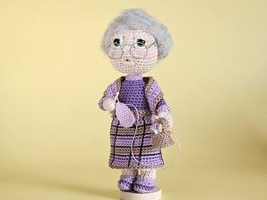 Gehäkelte Amigurumi-Oma mit grauem Haar, Brille, lila Kleidung, Häkelnadel und Garnkorb