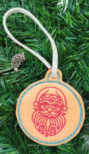 Runder Filzanhänger mit rot gesticktem Nikolausgesicht, türkisfarbener Rand und weißer Aufhängeschlaufe auf Tannengrün