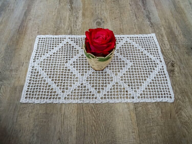 Weißes rechteckiges gehäkeltes Deckchen mit Gitter- und Rautenmuster auf Holz, rote Rose in kleinem Korb