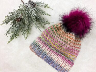Bunte Strickmütze mit Rippenbündchen und dunkelpinkem Bommel auf weißem, flauschigem Untergrund, daneben ein gefrosteter Tannenzweig