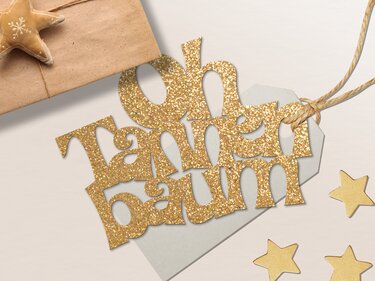 Selbstgemachter goldglitzernder 'Oh Tannenbaum'-Geschenkanhänger neben einem Päckchen und goldenen Sternen.