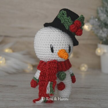 Gehäkelter Amigurumi‑Schneemann mit schwarzem Zylinder, rotem Schal und Karottennase vor hellem Holzhintergrund mit Lichterkette.