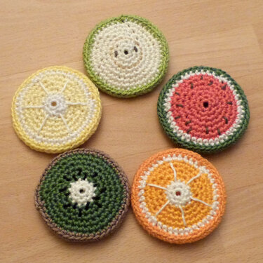 Fünf gehäkelte Untersetzer in Fruchtscheiben-Optik (Zitrone, Kiwi, Orange, Wassermelone, Limette) auf hellem Holz.