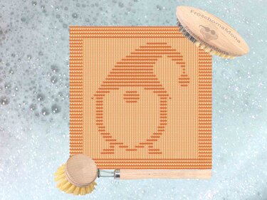 Gestricktes quadratisches Spültuch in Orangetönen mit stilisiertem Wichtelmotiv, auf hellem Untergrund neben Holzbürsten