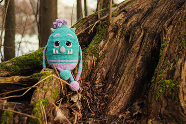 Gehäkeltes Amigurumi-Monster in Türkis und Lila auf moosbewachsenem Baumstamm