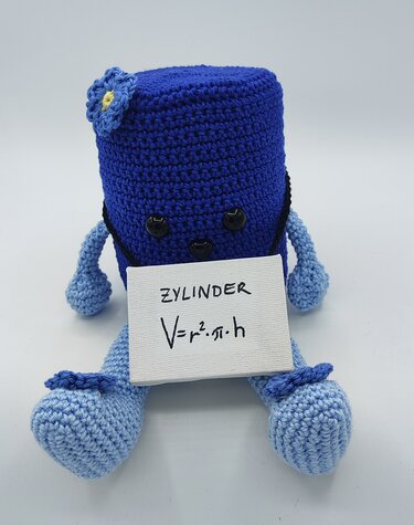 Blaues gehäkeltes Zylinder-Amigurumi mit hellblauen Gliedmaßen, kleiner Blume am Kopf und einem weißen Schild mit der Aufschrift 'ZYLINDER V=r²·π·h'.