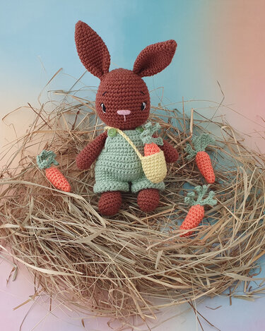 Brauner Amigurumi-Hase in grüner Latzhose mit gelber Umhängetasche sitzt im Strohnest, umgeben von kleinen Häkelmöhren.