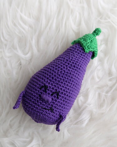 Lila gehäkelte Aubergine mit lächelndem Gesicht und grünem Stiel auf weißem flauschigem Stoff