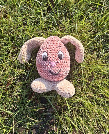 Pink egg-shaped amigurumi bunny with beige floppy ears and feet sitting on green grass