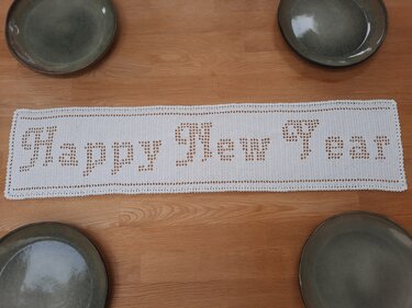 Weißer gehäkelter Filet-Tischläufer mit eingearbeitetem Schriftzug 'Happy New Year' auf Holztisch, umgeben von Tellern