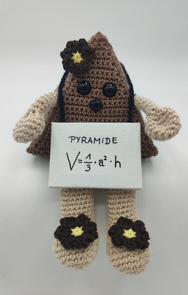 Pyramidenförmiges gehäkeltes Amigurumi in Braun und Beige mit Knopfaugen, Blumenapplikationen und Schild mit der Formel V = 1/3 · a² · h