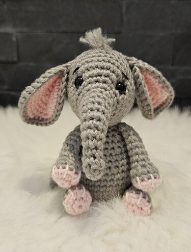 Gehäkelter grauer Amigurumi‑Elefant mit rosa Innenohren und rosa Füßen auf weißem Fell