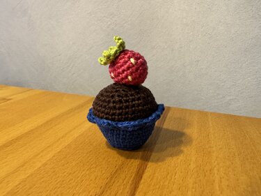 Gehäkelter Cupcake in blauem Förmchen mit brauner Kuppel und roter Erdbeere