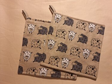 Zwei quadratische Topflappen aus beigem Stoff mit kleinem Schafsmuster und Aufhängeschlaufen