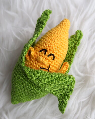 Gehäkelter gelber Maiskolben mit grüner Hülle und lächelndem Gesicht auf weißem Hintergrund.