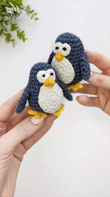 Two small dark blue and white crocheted amigurumi penguins with yellow beaks and feet, held in hands against a white background.