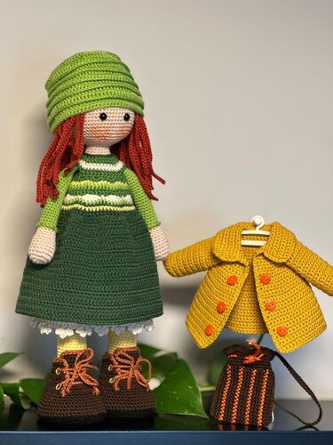 Crochet amigurumi doll with red hair, green hat and green dress, standing next to a yellow crochet coat on a hanger and a small striped bag