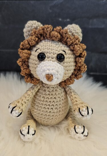 Beige gehäkelter Amigurumi-Löwe mit brauner lockiger Mähne sitzend auf hellem Fell