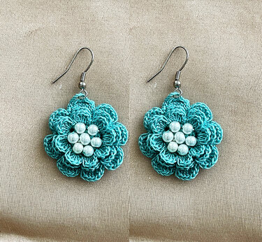 Pair of teal crocheted flower earrings with white pearl beads and silver-tone hooks on beige fabric.