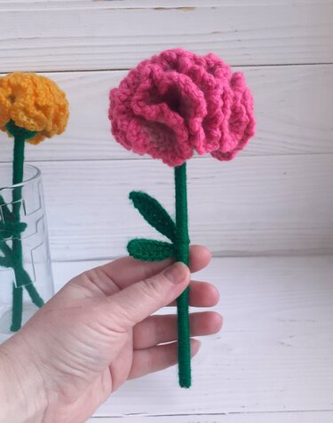 Hand holding a pink crocheted flower with a wrapped green stem and two small green leaves; a yellow crocheted flower in a glass vase is blurred behind.
