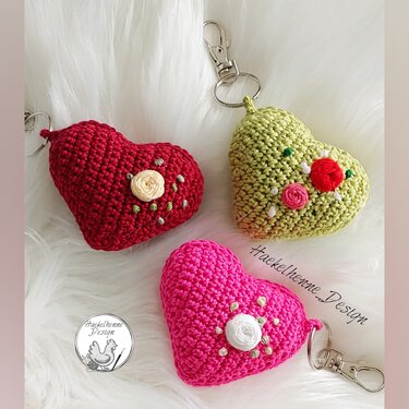 Drei gehäkelte Herz-Schlüsselanhänger in Rot, Grün und Pink mit kleinen Blüten und Metall-Karabiner auf weißem, flauschigem Untergrund.