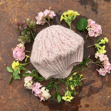 Rosafarbene, gestrickte Damenmütze mit auffälligem Strukturmuster, flach auf rostigem Untergrund platziert und von rosa und gelben Blumen umgeben.