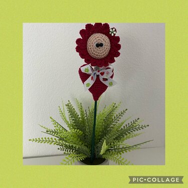 Rote gehäkelte Amigurumi-Blume mit beigem Zentrum und zwei schwarzen Knopfaugen, rotes Herz mit Schleife in einem Topf mit künstlichen Blättern.