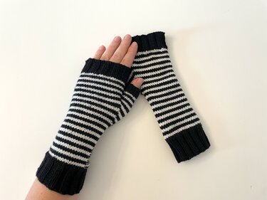 Hand trägt schwarz-weiß gestreifte Armstulpe, daneben liegt die zweite Stulpe flach auf hellem Hintergrund.