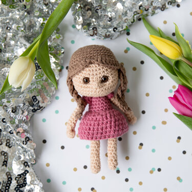 Gehäkelte Amigurumi-Puppe mit rosa Kleid und geflochtenen Zöpfen neben Tulpen