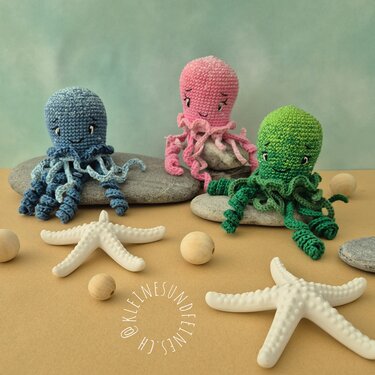 Drei gehäkelte Amigurumi‑Kraken in Blau, Rosa und Grün mit spiraligen Tentakeln auf Steinen