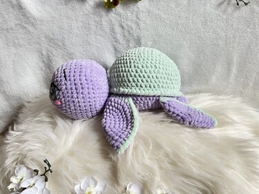 Gehäkelte Amigurumi‑Schildkröte in Pastelllila und Mintgrün auf weißem Fell