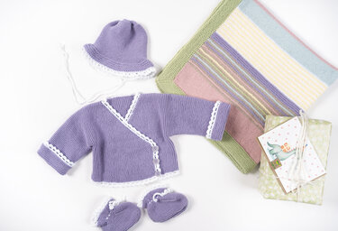 Lavendelfarbene Wickeljacke, Mütze und Schühchen mit weißer Kante neben einer pastellgestreiften Babydecke und einem verpackten Geschenk mit Karte.