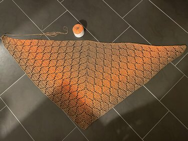 Gehäkeltes Dreieckstuch in Orange- und Brauntönen mit Spitzenmuster, daneben ein orangefarbenes Garnknäuel auf dunklem Fliesenboden