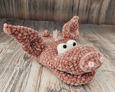 Rosa gehäkeltes Amigurumi in Schweineform mit großen Ohren und aufgesetzter Schnauze, liegend auf Holzboden