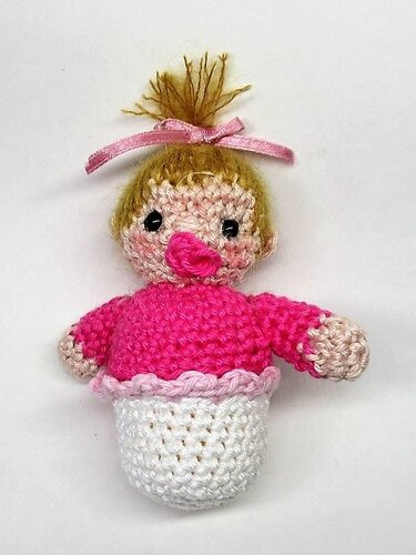 Gehäkeltes Amigurumi-Baby mit rosa Oberteil und weißem Unterteil, blondem Haarschopf, rosa Schleife und pinkem Schnuller