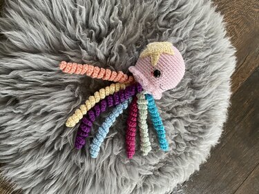 Handgehäkelte rosafarbene Amigurumi-Qualle mit mehrfarbigen spiralförmigen Tentakeln auf grauem, flauschigem Untergrund.