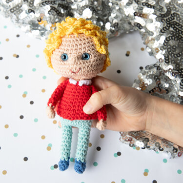 Hand hält gehäkelte Amigurumi-Puppe mit blonden Locken, rotem Pullover und hellblauer Hose