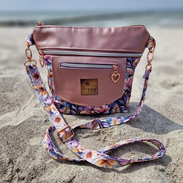 Rosafarbene Umhängetasche in Lederoptik mit Blumenstoff, zwei Reißverschlüssen und Herzanhängern im Sand am Strand.