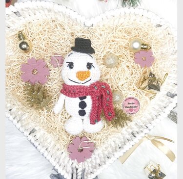 Handgehäkelter Schneemann mit rotem Schal und Karottennase in einer herzförmigen Schale mit Holzwolle und Weihnachtsdekoration.