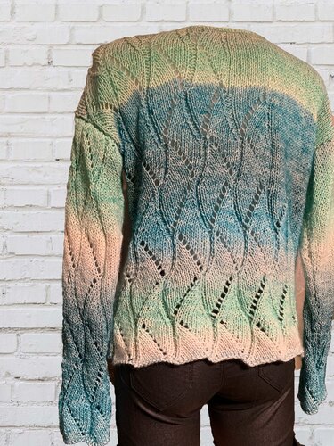 Nahaufnahme der Rückseite eines gestrickten Damenpullovers mit Blatt-Ajourmuster und Ombré-Farbverlauf in Creme, Blau und Grün vor weißer Ziegelwand.