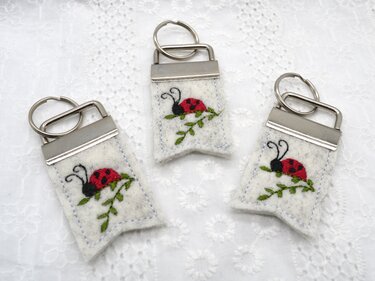 Drei helle Filz-Schlüsselanhänger mit gesticktem roten Marienkäfer, Metallklemmen und Schlüsselringen auf weißer Stoffunterlage