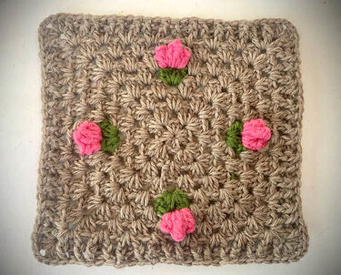 Beiges Granny Square mit vier applizierten rosa Rosen und grünen Blättern