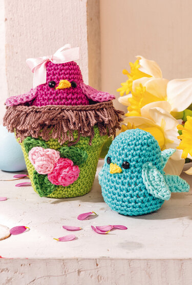 Zwei gehäkelte Amigurumi-Vögel: ein rosa Vogel mit Schleife im grünen, blumengeschmückten Nest, daneben ein türkisblauer Vogel vor Narzissen.