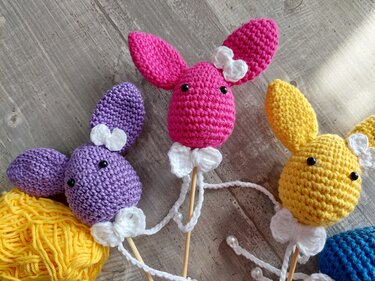 Drei gehäkelte Hasenköpfe in Pink, Lila und Gelb mit weißen Schleifen auf Holzstäben