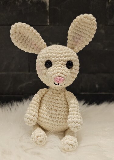 Gehäkelter cremefarbener Amigurumi-Hase mit rosa Nase und schwarzen Augen auf weißem Fell.