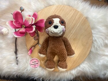 Handgehäkeltes Faultier-Amigurumi in Braun und Weiß mit rosa Bäckchen auf rundem Holzbrett