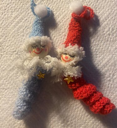 Zwei gehäkelte Wichtel, blau und rot, mit bemalten Holzgesichtern, flauschigen Krägen und goldenen Sternen.