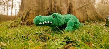 Grünes gehäkeltes Krokodil-Amigurumi mit weißen Zähnen im Gras vor einem Baumstamm.