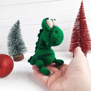 Small green crocheted dinosaur amigurumi with large eyes and back spikes sitting in an open hand, with miniature festive trees and a red bauble on a white surface.